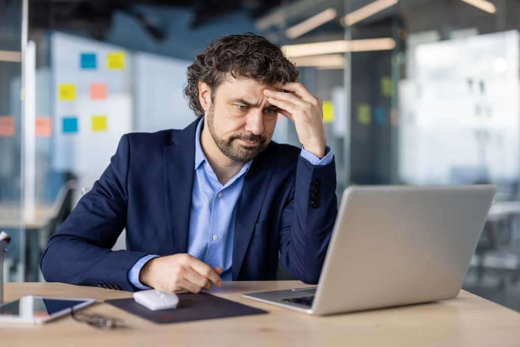 Businessman experiencing stress working on laptop in office. Professional looks frustrated and worried, dealing with issue. Man in suit facing pressure at work. Implements phone and tablet on desk.