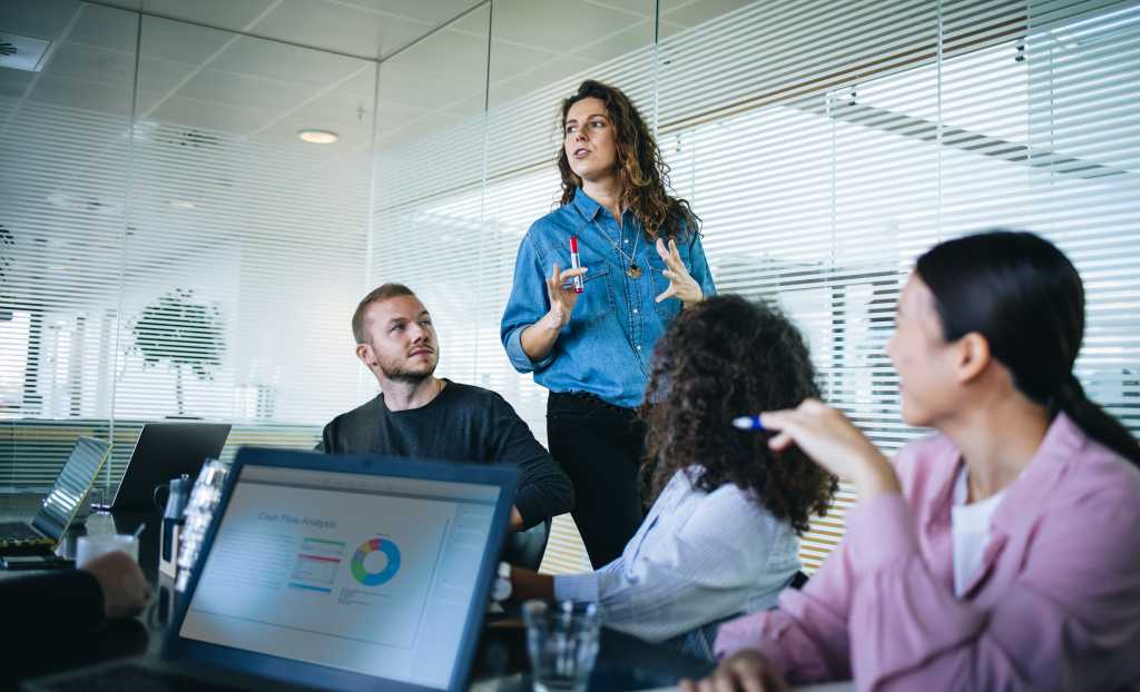 Businesswoman walking in office boardroom and sharing a strategy with team sitting at table. Female manager explaining new business plan with coworkers sitting at conference table in office.