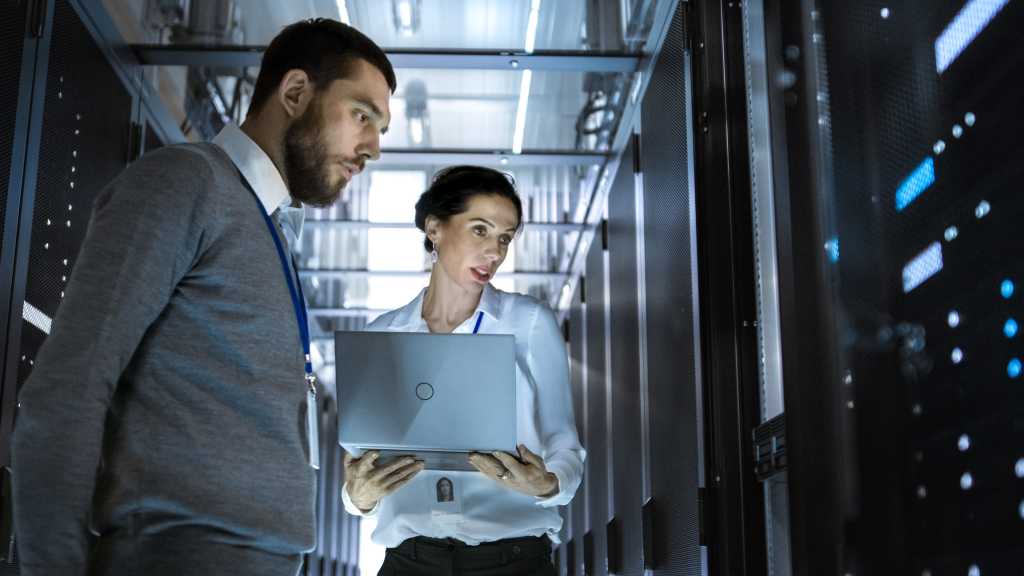 Male Server Engineer is Looking at Server Rack Cabinet. His Female Colleague Works on a Laptop Beside Him. They're in Big Data Center.